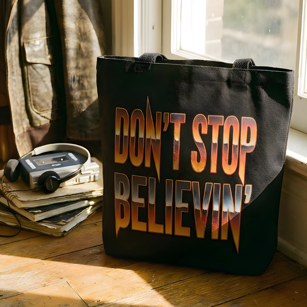 Black tote bag with 'Don't Stop Believin'' text on a wooden floor with a window in the background.