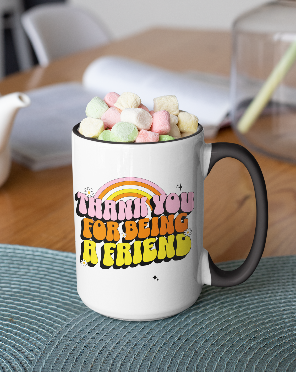 Mug with colorful text and marshmallows on a table with a teapot and plant in the background