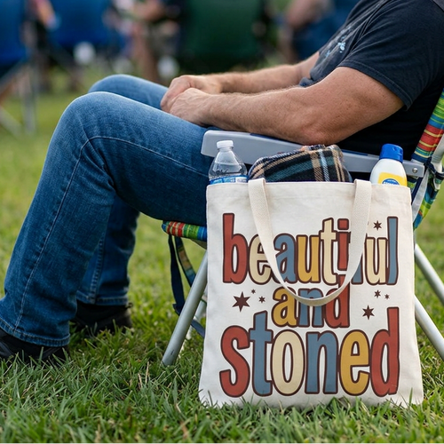 A man sits in a folding lawn chair on a grassy outdoor area, with a cream canvas tote bag placed in front of the chair. The tote bag has colorful text reading 'beautiful and Stoned' with small star accents.