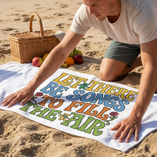 A man kneels on a sandy beach next to the ocean, smoothing out a white towel printed with the colorful retro-style text 'LET THERE BE SONGS TO FILL THE AIR'. A wicker picnic basket, fruit, and a vintage radio are nearby.