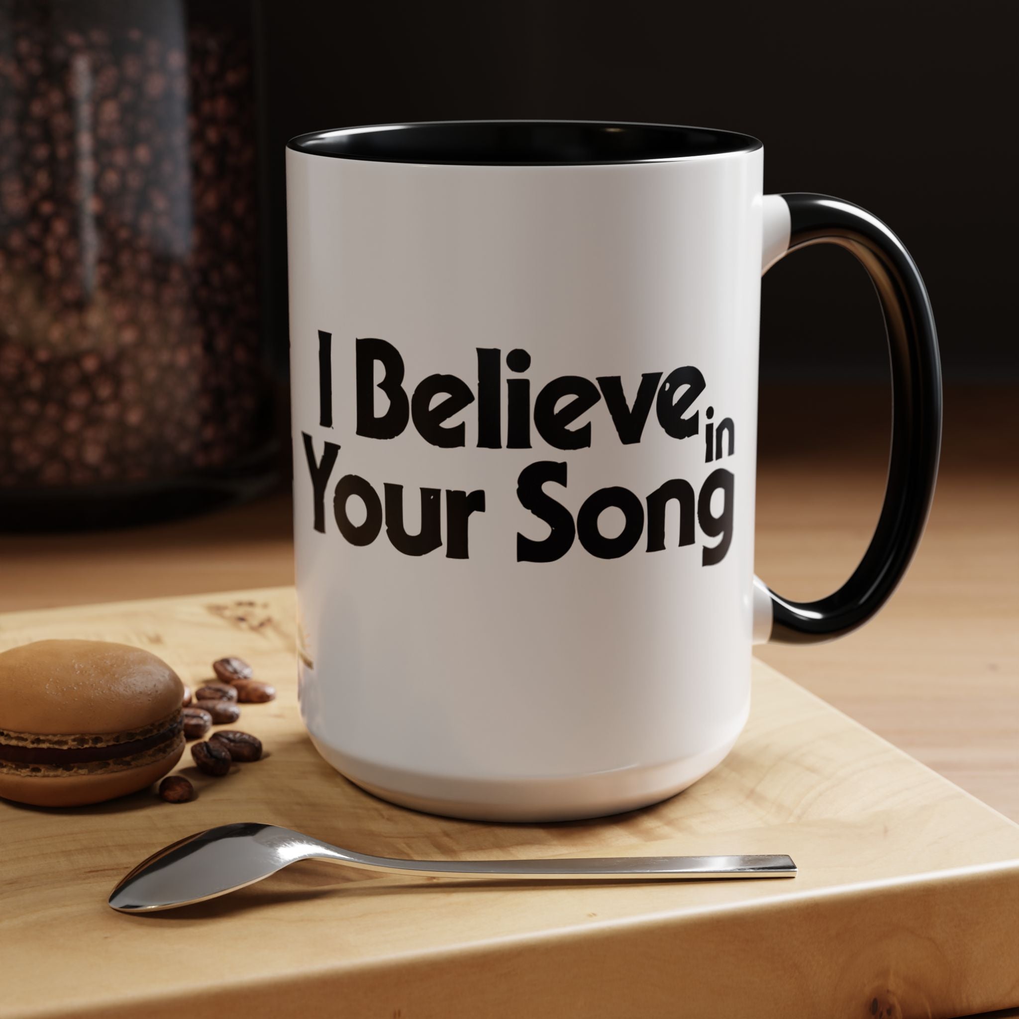 White mug with 'I Believe in Your Song' text on a wooden surface with coffee beans and a spoon.