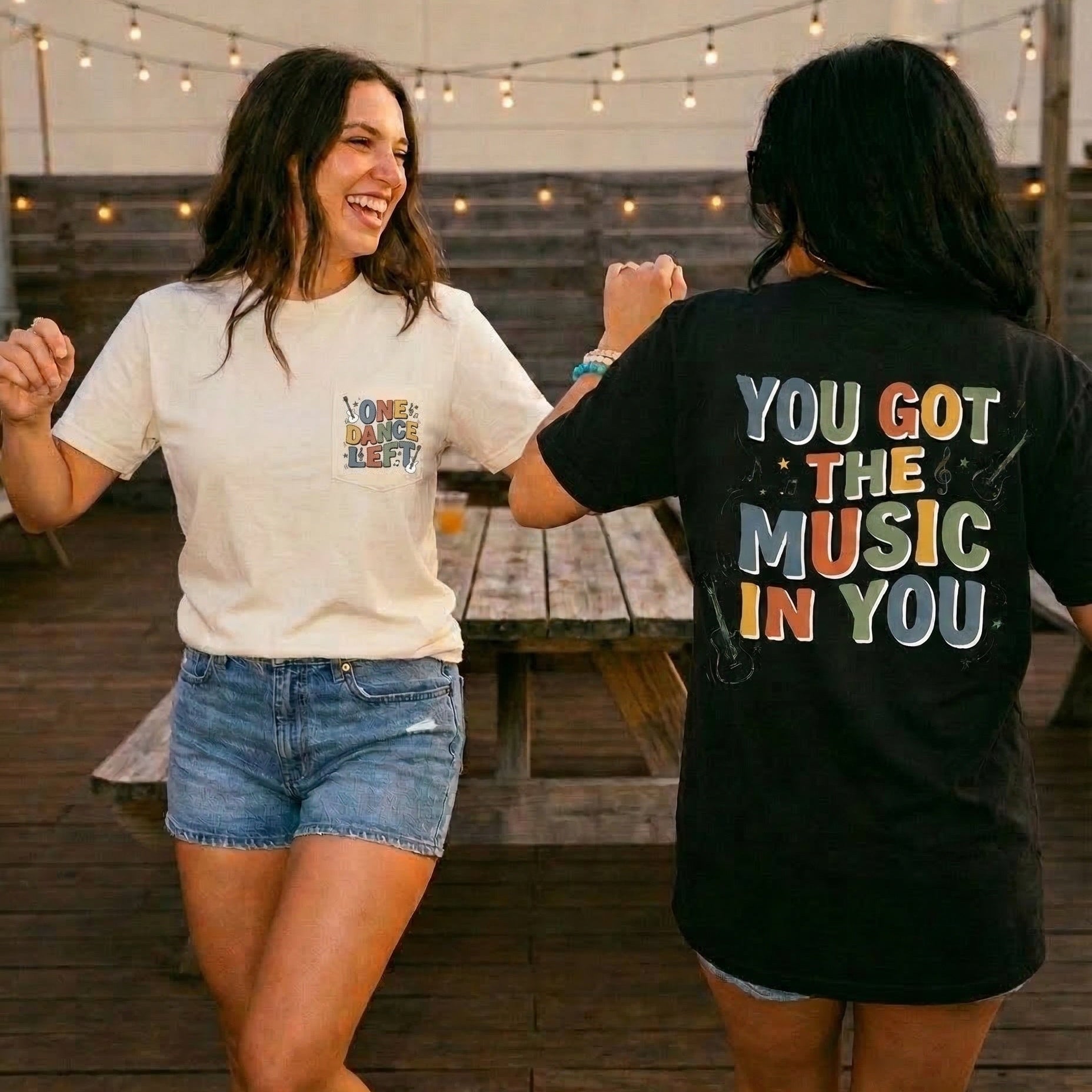 Two people dancing outdoors with one wearing a white t-shirt and denim shorts, and the other wearing a black t-shirt with colorful text.