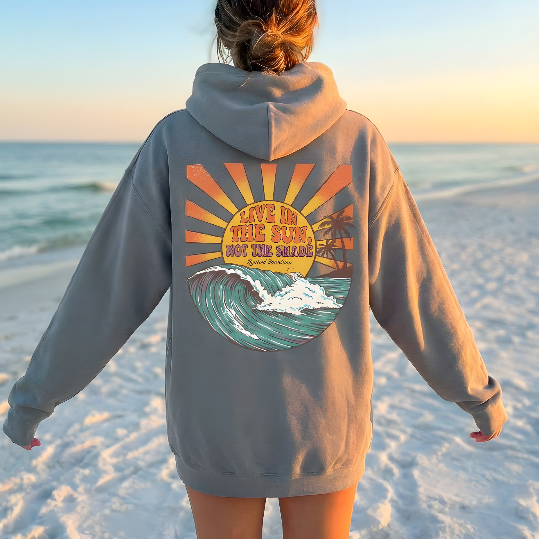 Person wearing a hoodie with a beach-themed design on a sandy beach at sunset.
