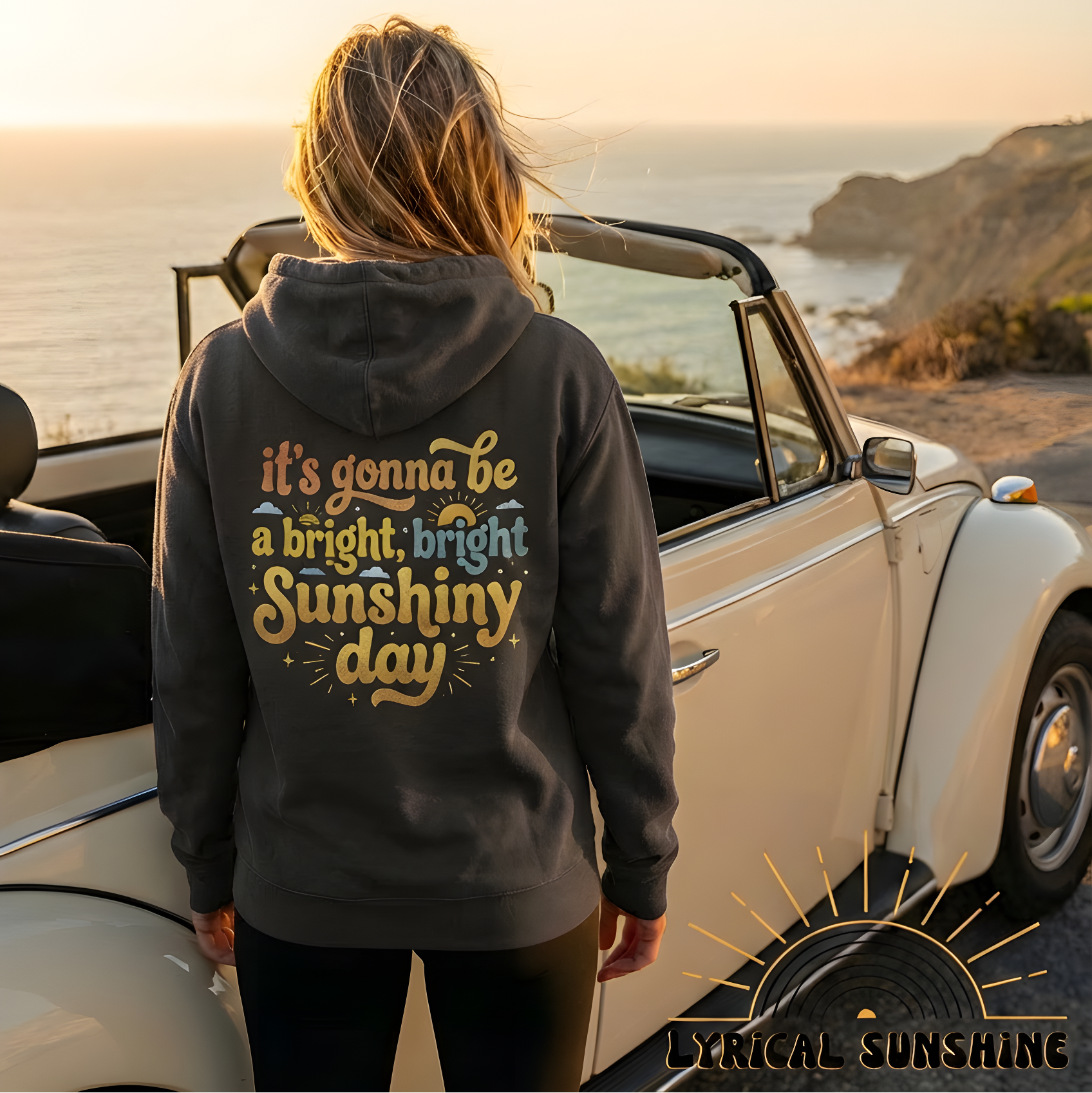 Person wearing a hoodie with a motivational quote, standing next to a vintage car by the ocean at sunset.