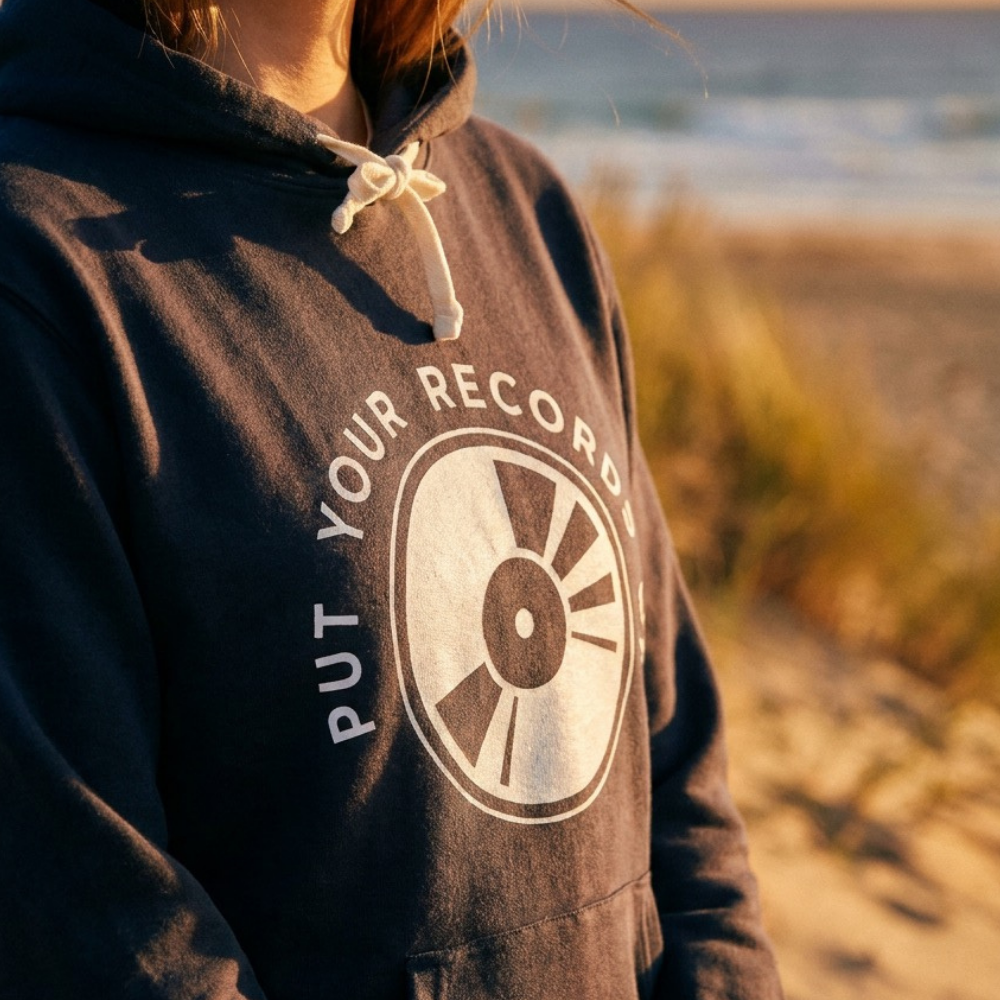 Person wearing a hoodie with 'Put Your Records' design on a beach at sunset.