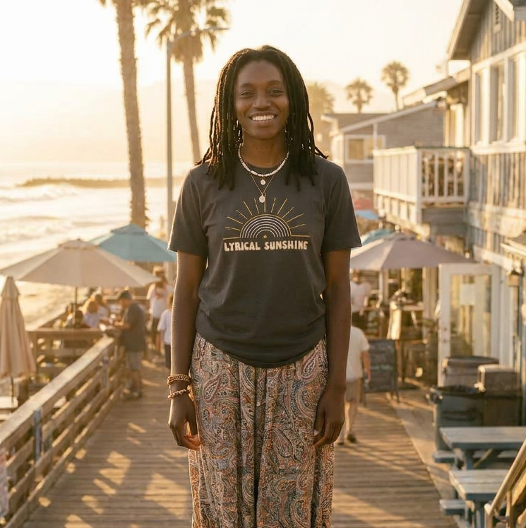 Woman standing on a wooden boardwalk by the beach with palm trees and houses in the background.