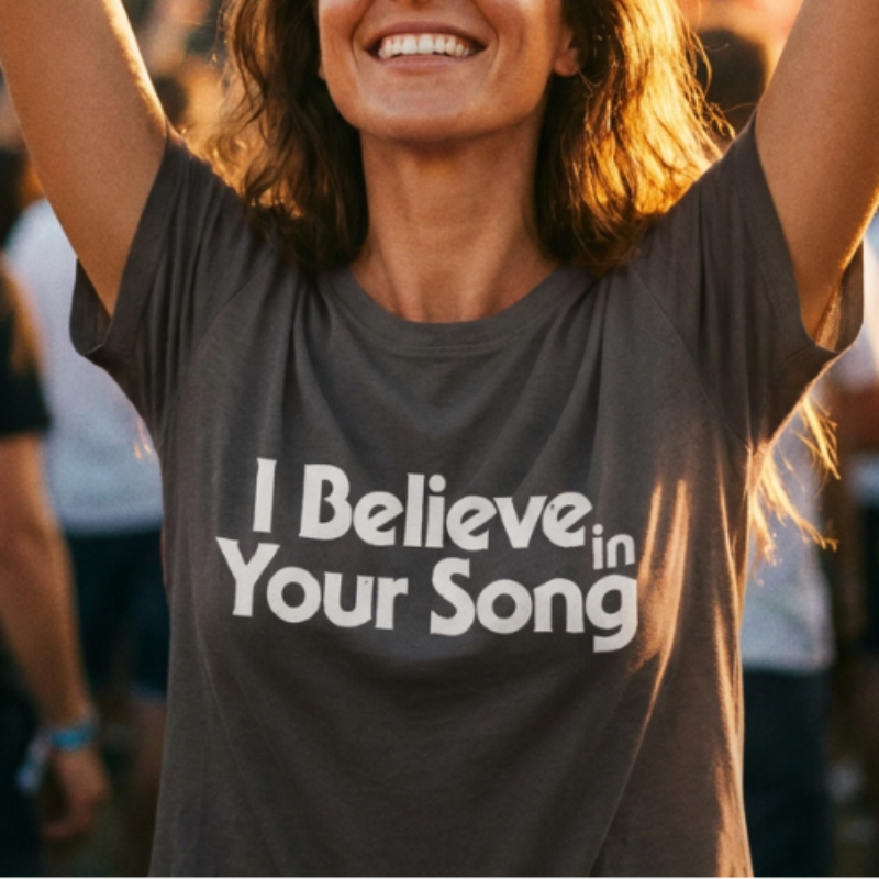 Woman at a concert wearing a t-shirt with 'I Believe in Your Song' text, arms raised.