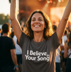 Woman at a concert wearing a t-shirt with 'I Believe in Your Song' text, arms raised.