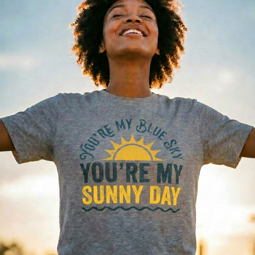 Person wearing a gray t-shirt with a sun graphic and text, standing outdoors with a blurred background