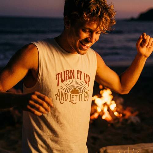 Man wearing a tank top with text by a campfire on a beach at sunset