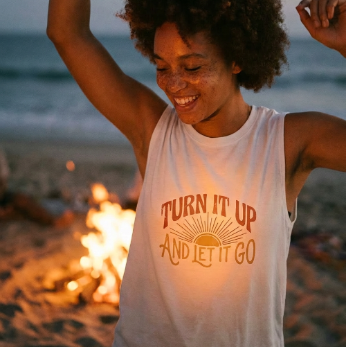 Person on a beach with a bonfire, wearing a tank top with text, at sunset.