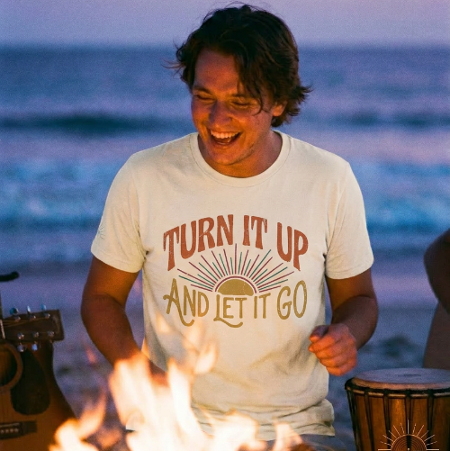 Person wearing a 'Turn It Up and Let It Go' shirt by a fire on a beach with ocean in the background