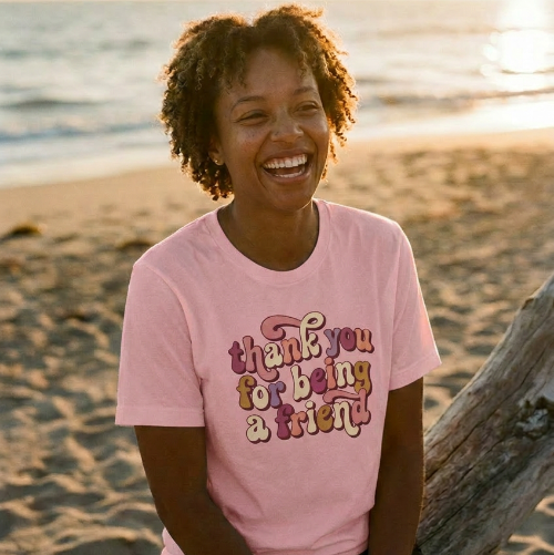 Person wearing a pink t-shirt with a message, sitting on a beach at sunset.