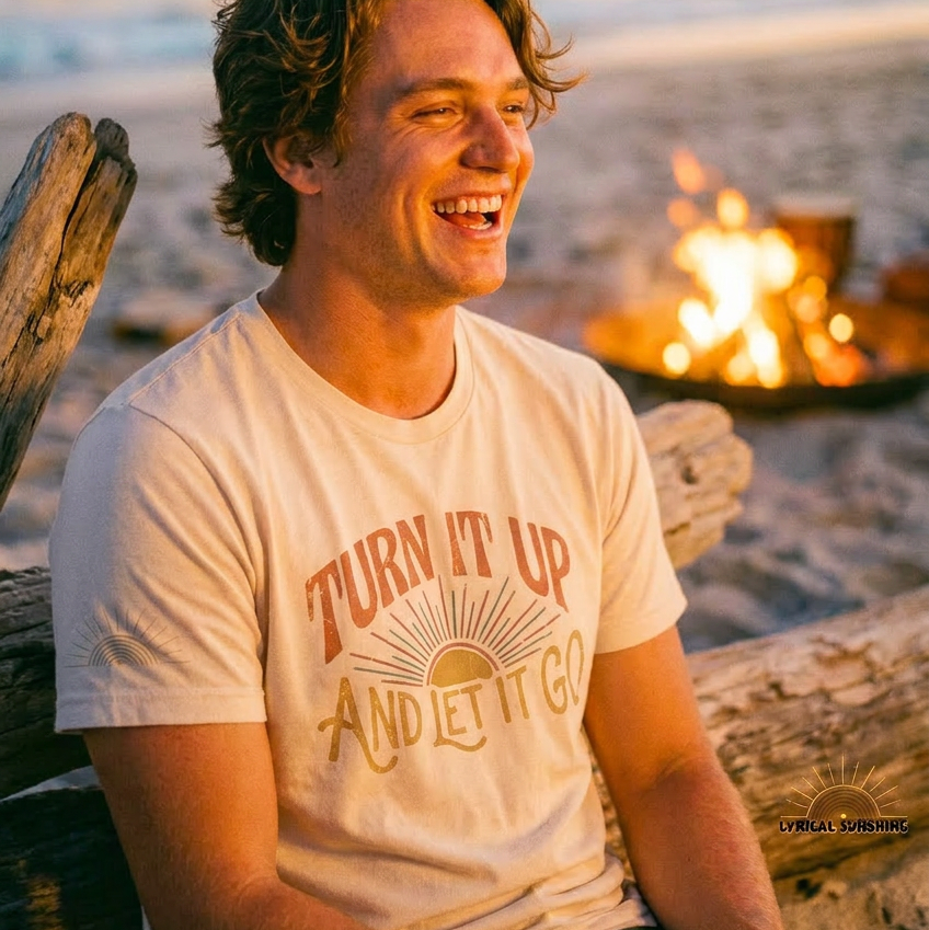 Man sitting by a campfire on a beach with 'Turn It Up And Let It Go' t-shirt.