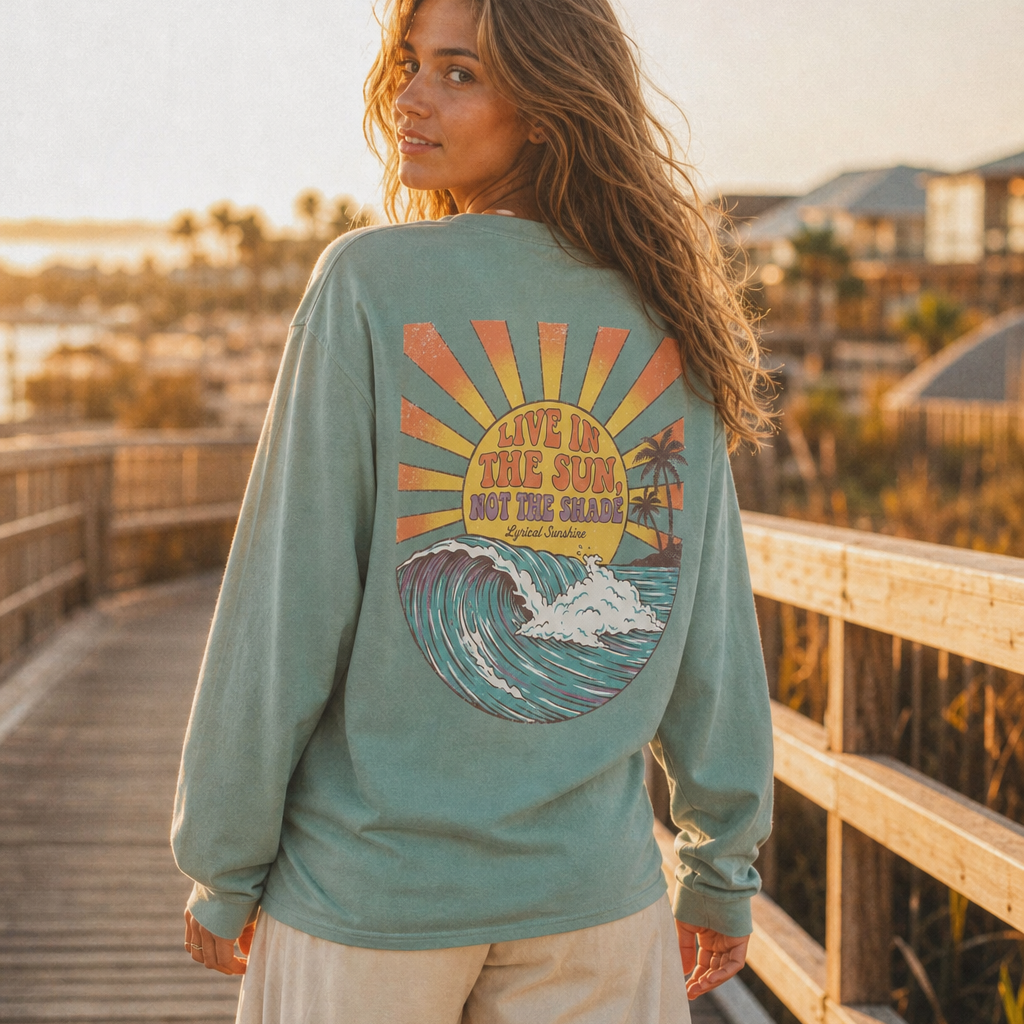 A woman stands on a wooden boardwalk at sunset wearing a sage green long-sleeve graphic t-shirt with a large retro wave design on the back, paired with cream pants.