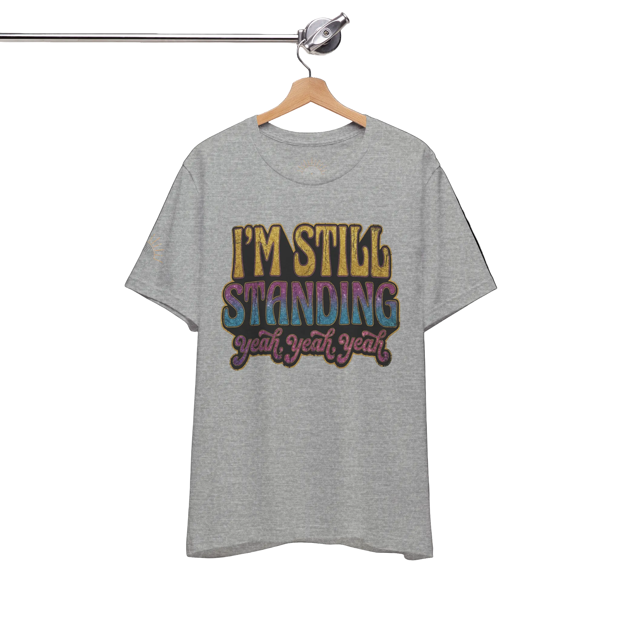 Gray t-shirt with colorful text 'I'M STILL STANDING yeah yeah yeah' on a hanger against a white background