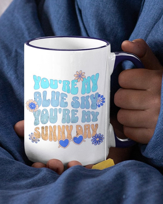 Person holding a mug with 'You're my blue sky, you're my sunny day' text, wrapped in a blue blanket.