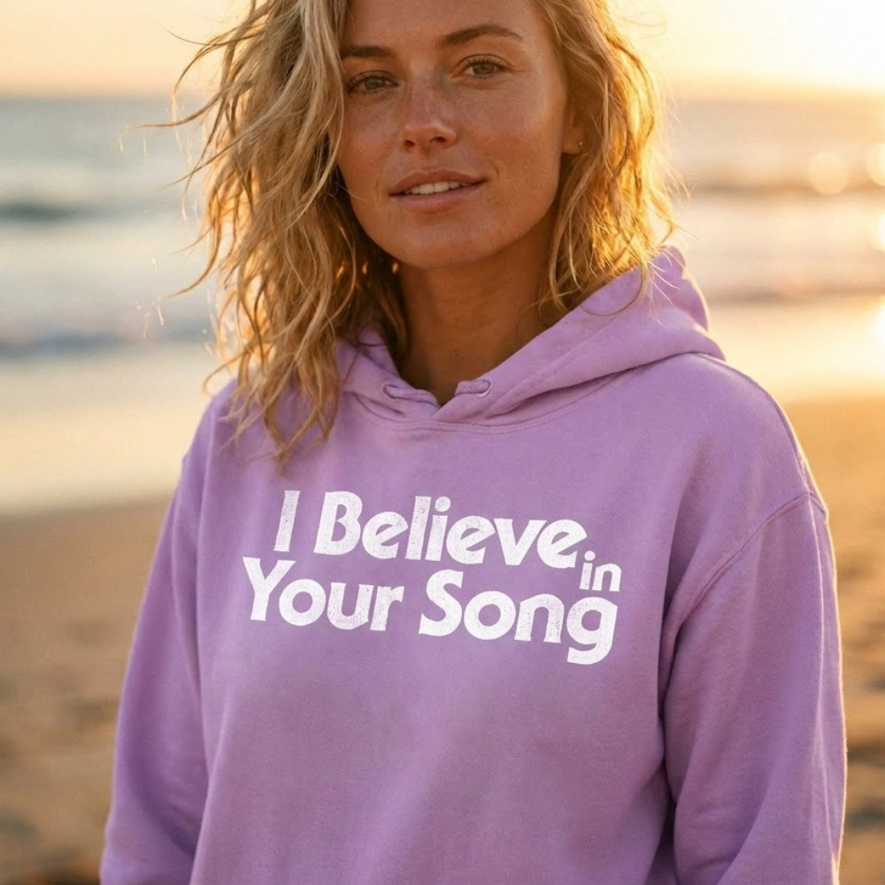 Woman wearing a purple hoodie with 'I Believe in Your Song' text at sunset.