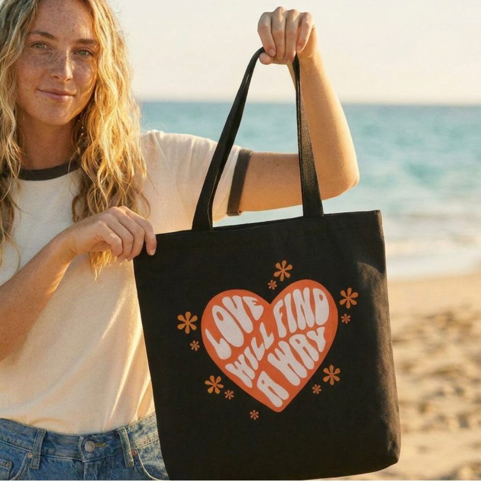 Woman holding a black tote bag with a heart design on a beach, with 'Lyrical Sunshine' logo in the corner.