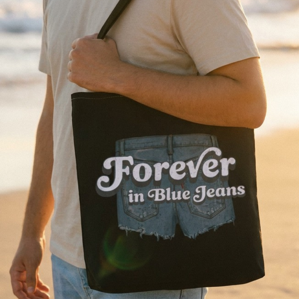 Person holding a tote bag with 'Forever in Blue Jeans' text on a beach.