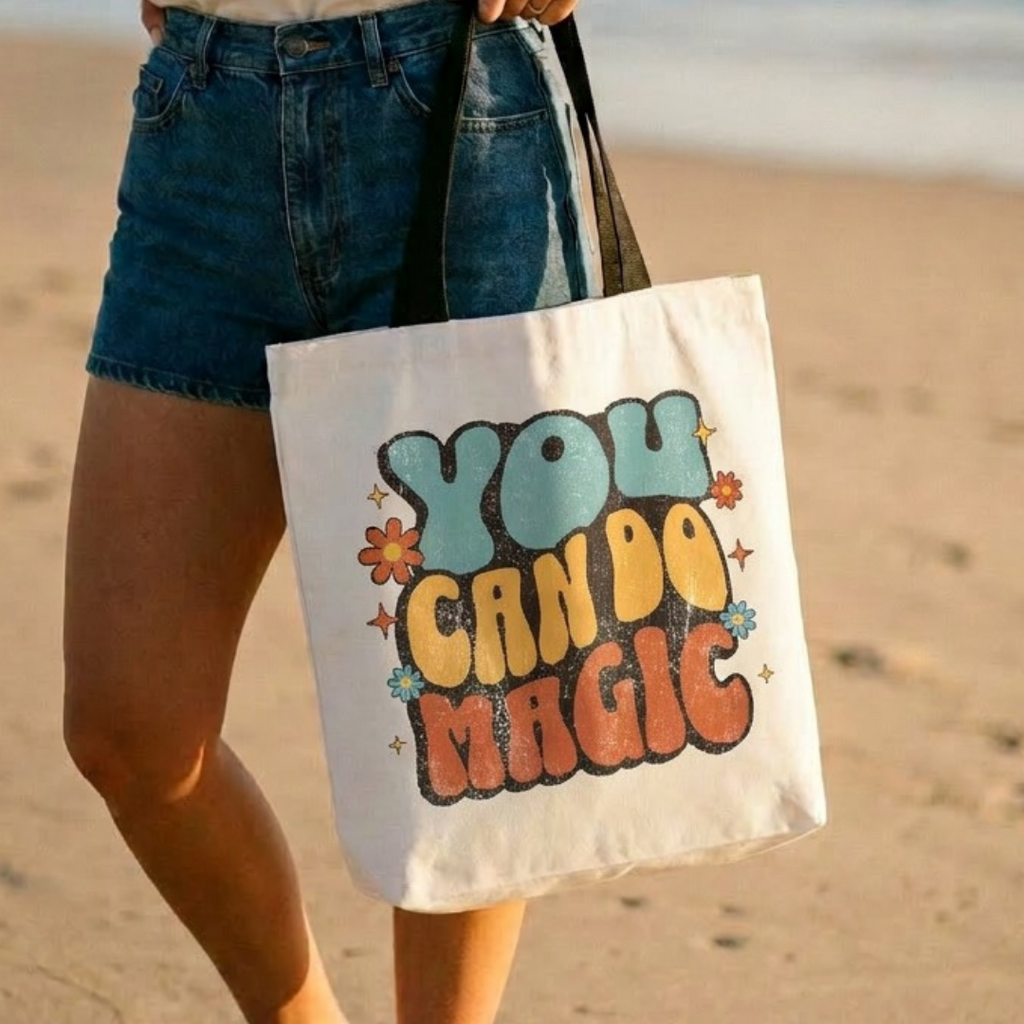 Person holding a tote bag with 'You Can Do Magic' text on a beach