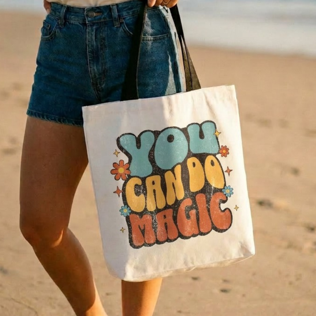 Person holding a tote bag with 'You Can Do Magic' text on a beach