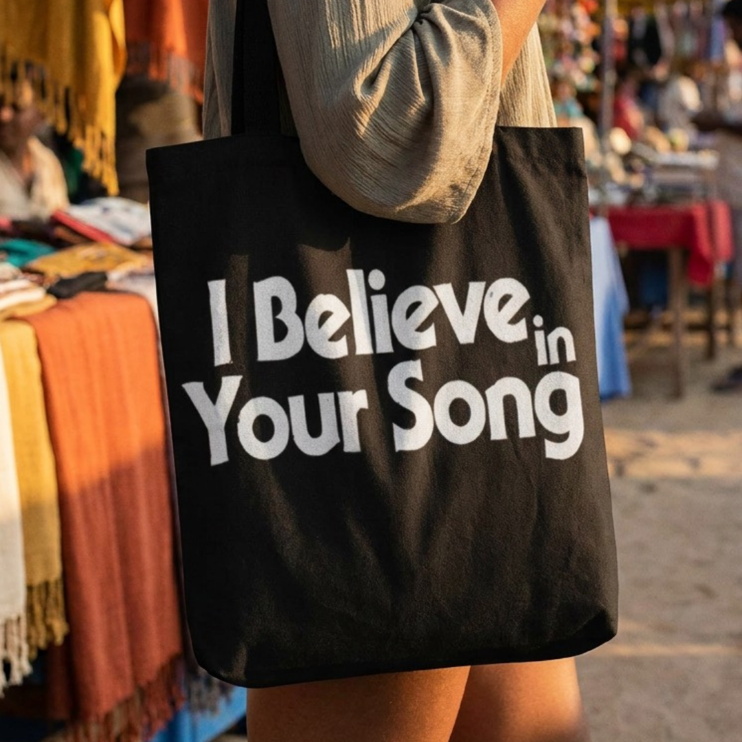 Person holding a black tote bag with 'I Believe in Your Song' text at an outdoor market.