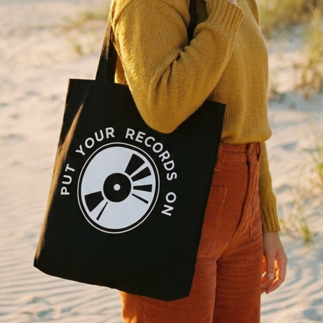 Person holding a black tote bag with a record player graphic and text on a beach.