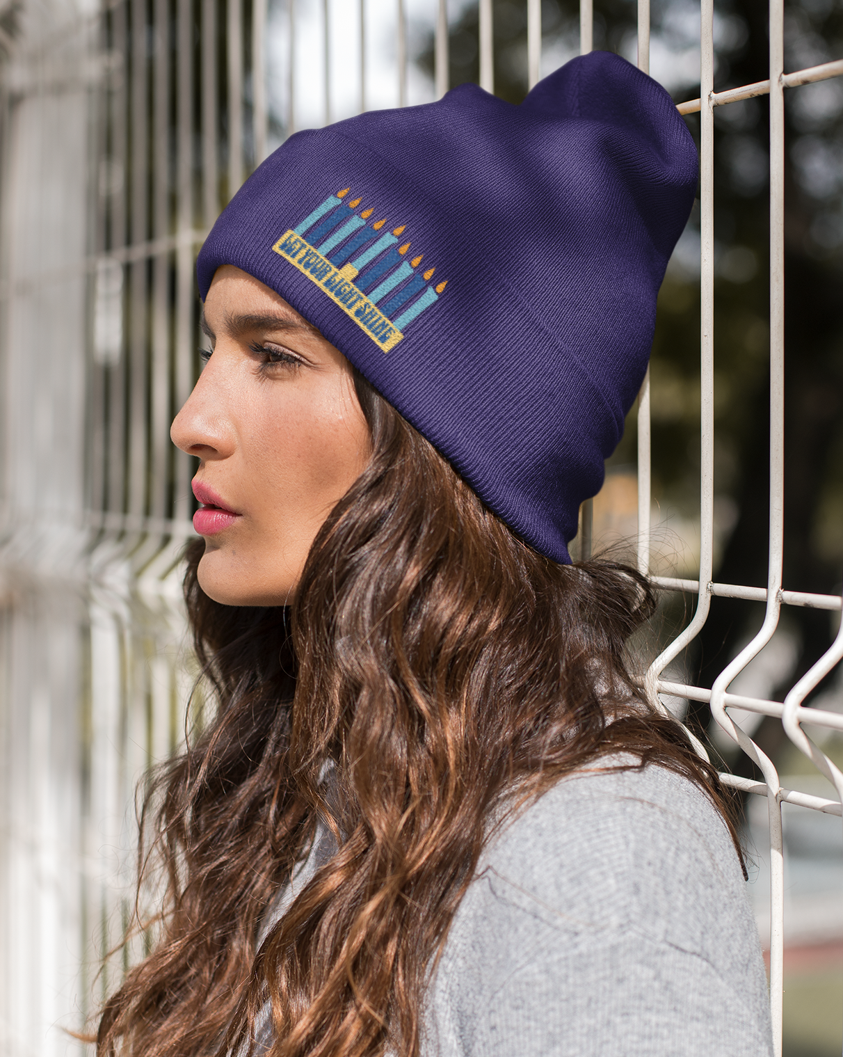 Woman wearing a purple beanie with a design, standing against a wire fence.