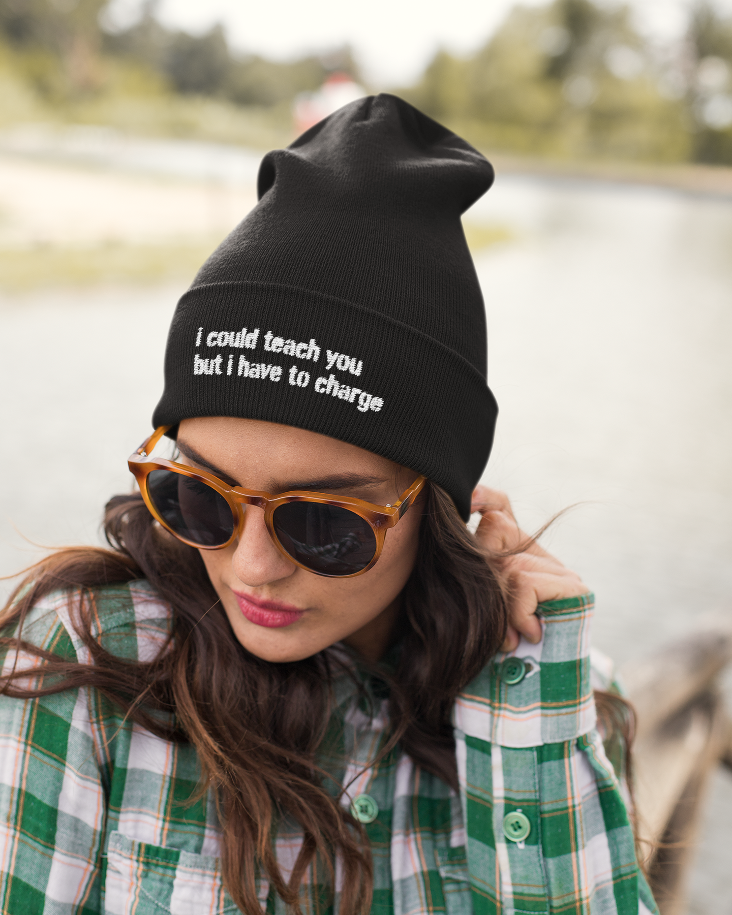 Person wearing a black beanie with text, sunglasses, and a green plaid shirt by a body of water.