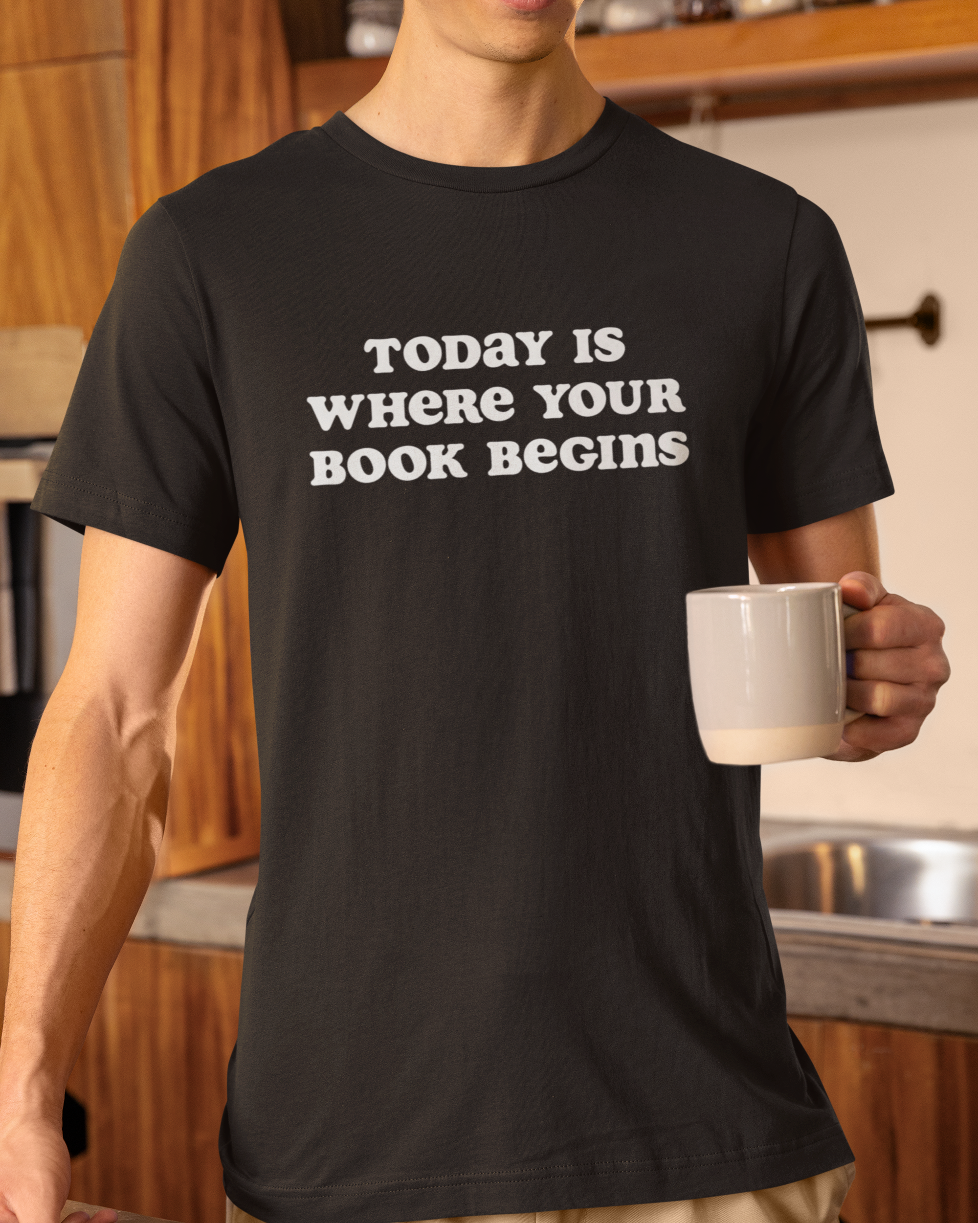 Person wearing a black t-shirt with 'Today is where your book begins' text, holding a mug in a kitchen.