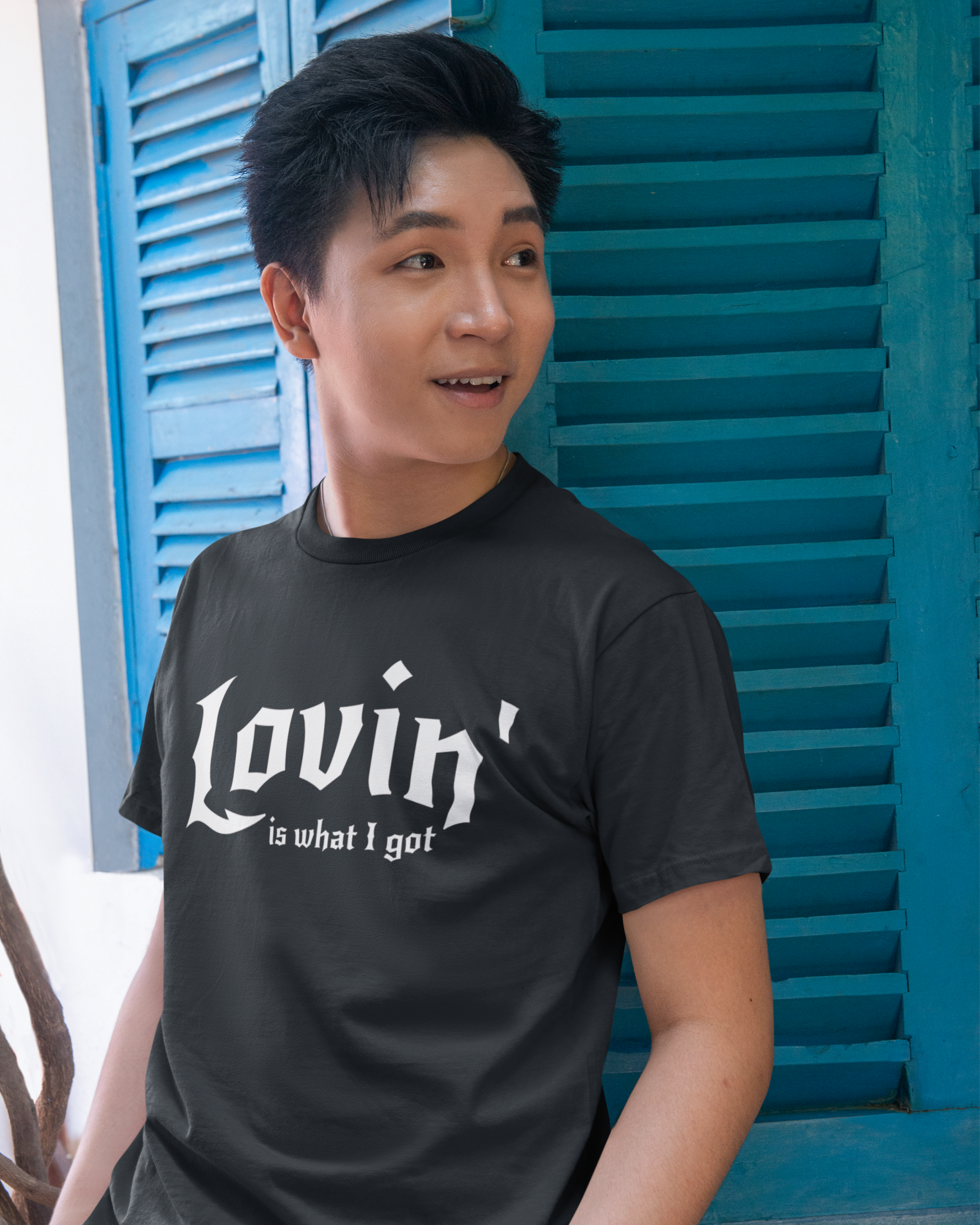 Person wearing a black t-shirt with 'Lovin is what I got' text in front of blue shutters.