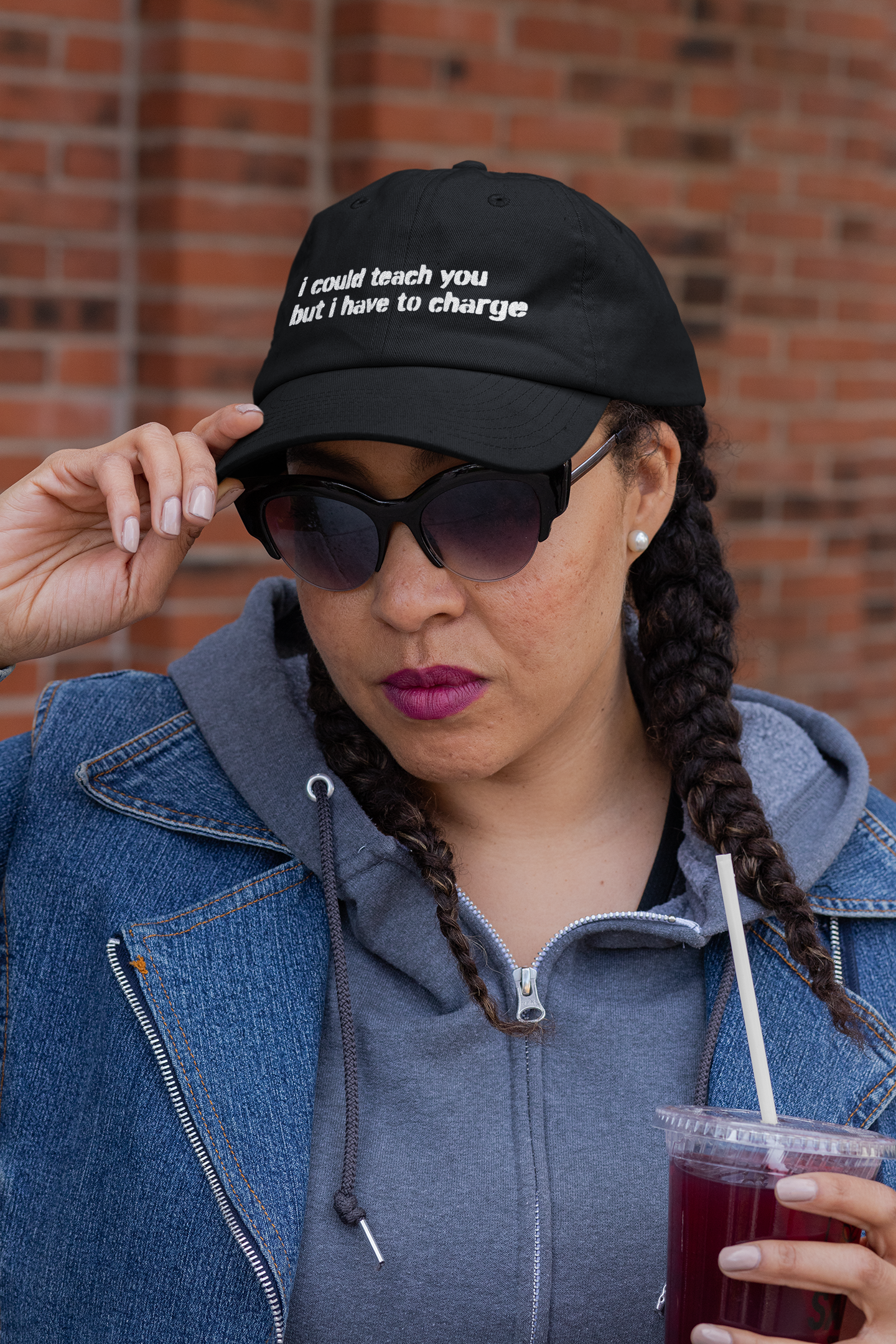 Woman wearing a black cap with text, sunglasses, and holding a drink against a brick wall.