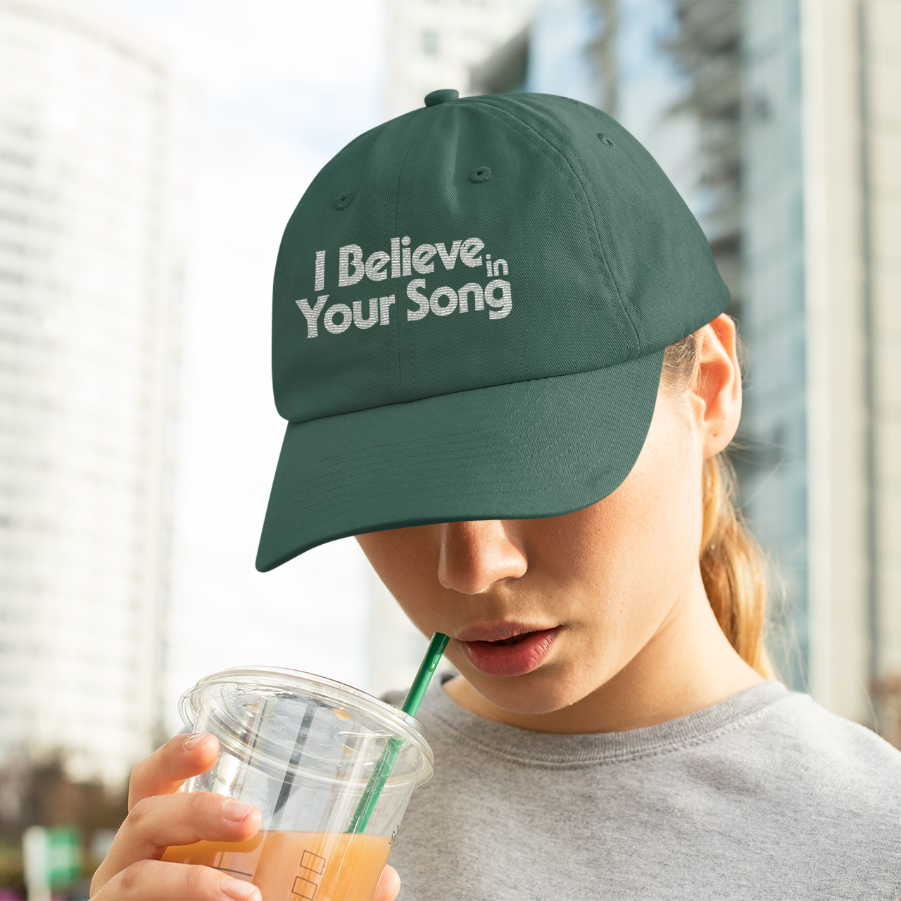 Person wearing a green cap with 'I Believe in Your Song' text, drinking from a straw.