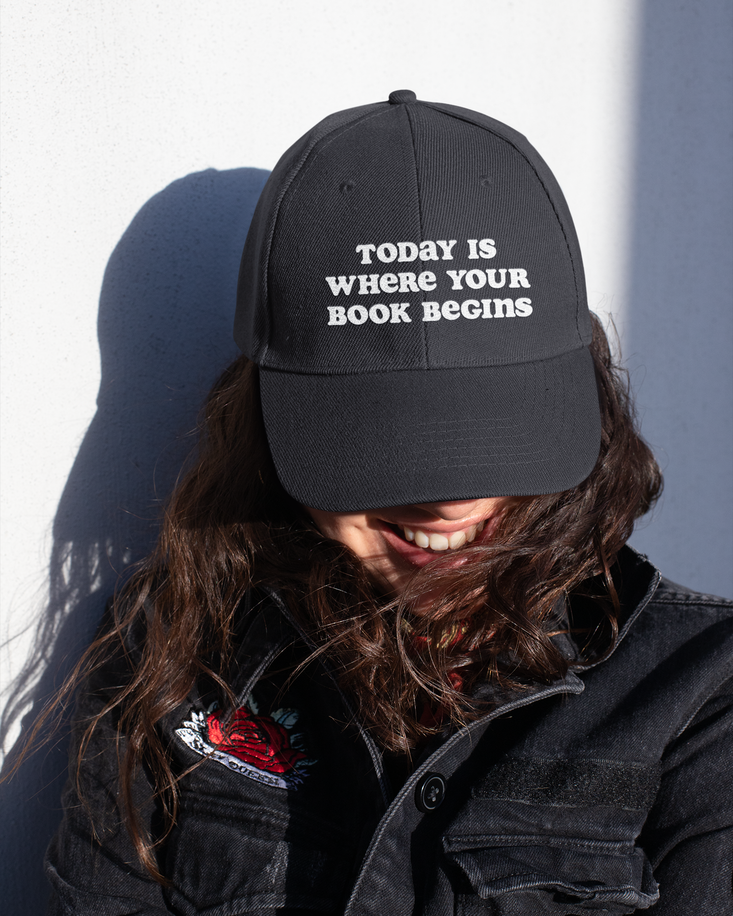 Person wearing a black cap with 'TODAY IS WHERE YOUR BOOK BEGINS' text, sitting against a white wall.