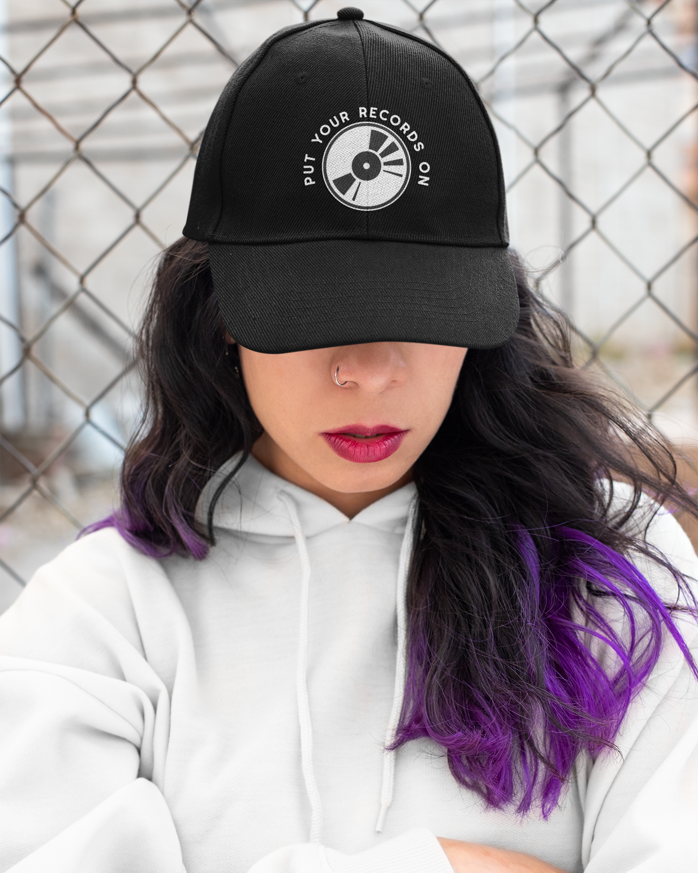 Person wearing a black cap with a logo and text, standing against a chain-link fence.