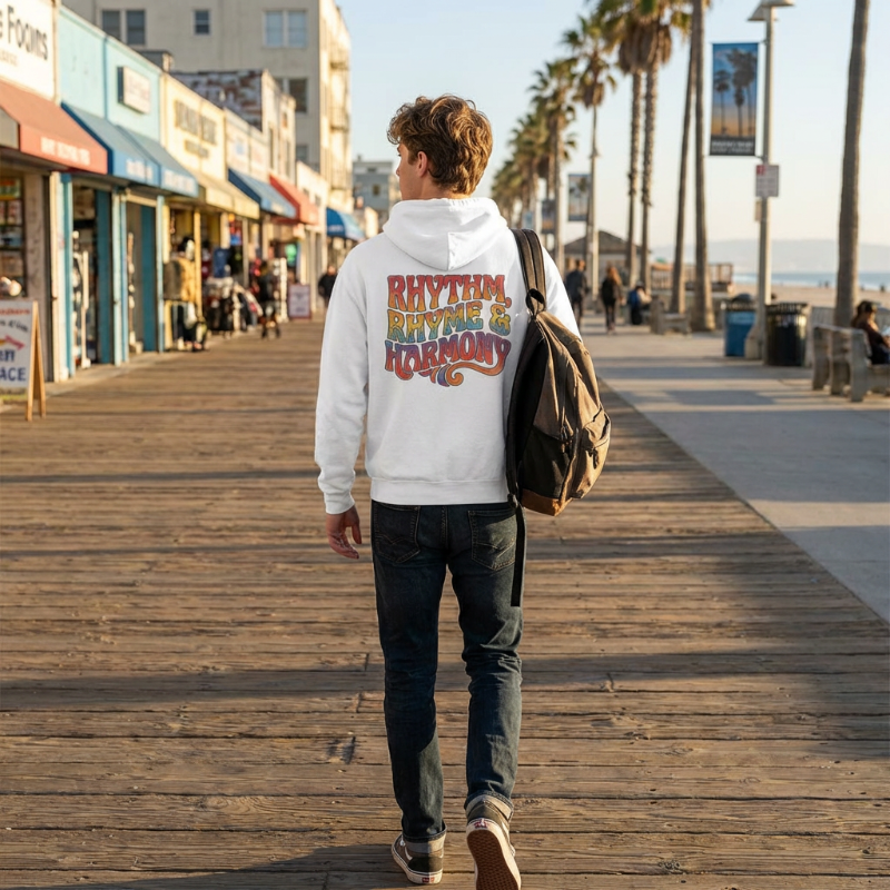 Person walking on a boardwalk wearing a hoodie with colorful text, palm trees and buildings in the background.