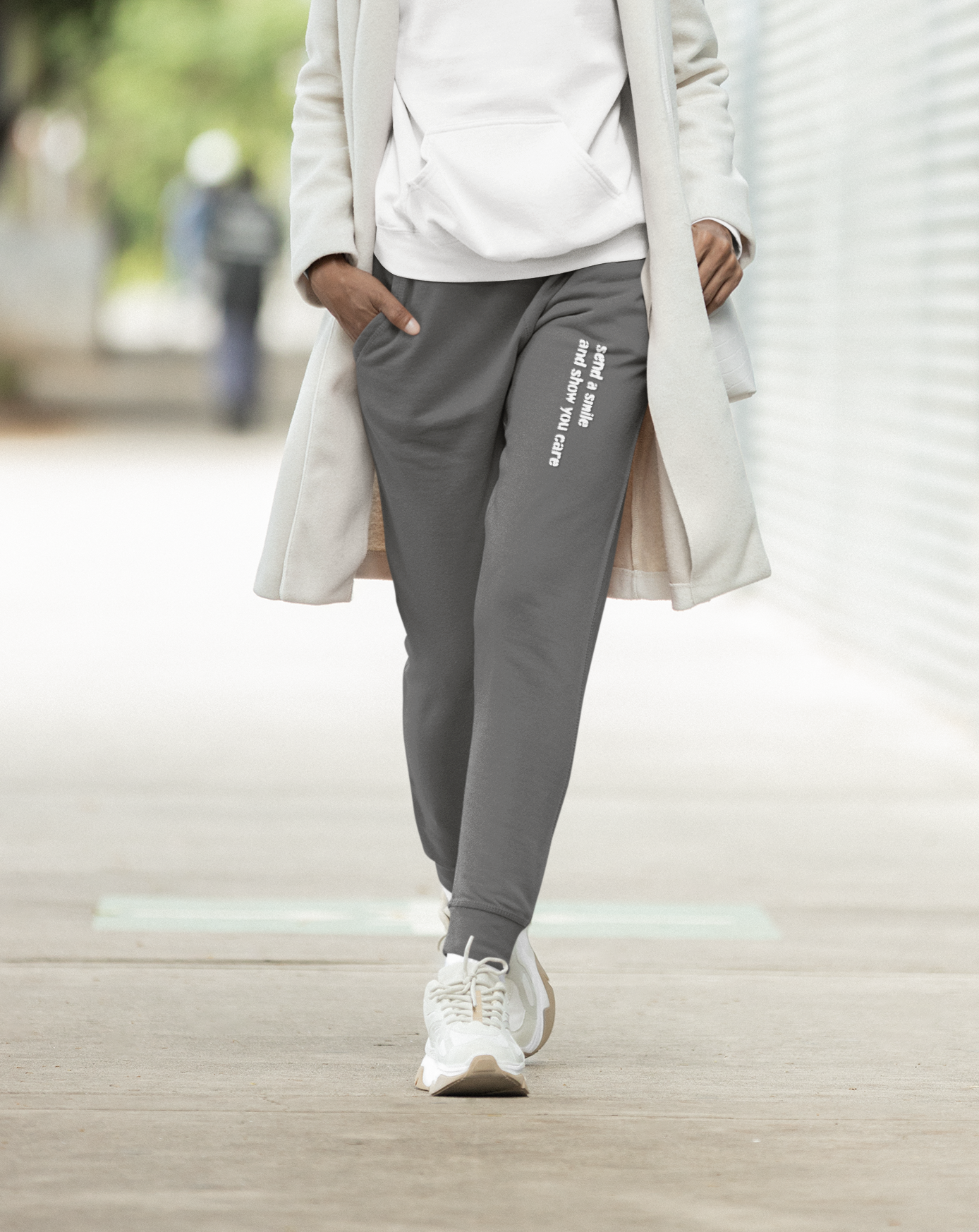 Person wearing a white hoodie, gray jogger pants, and a long beige coat walking on a sidewalk.