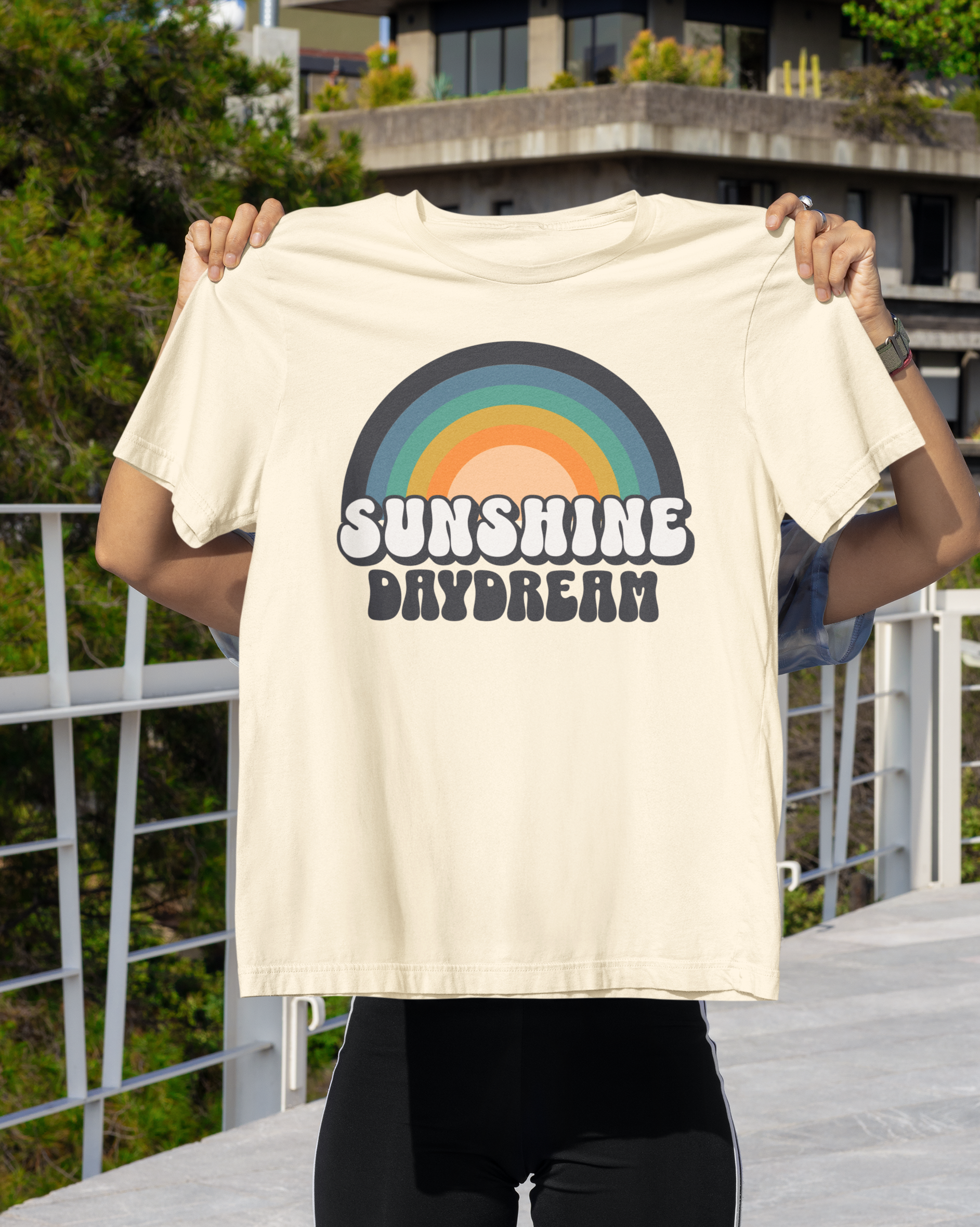 Person holding a t-shirt with a rainbow design and 'Sunshine Daydream' text against a scenic background.