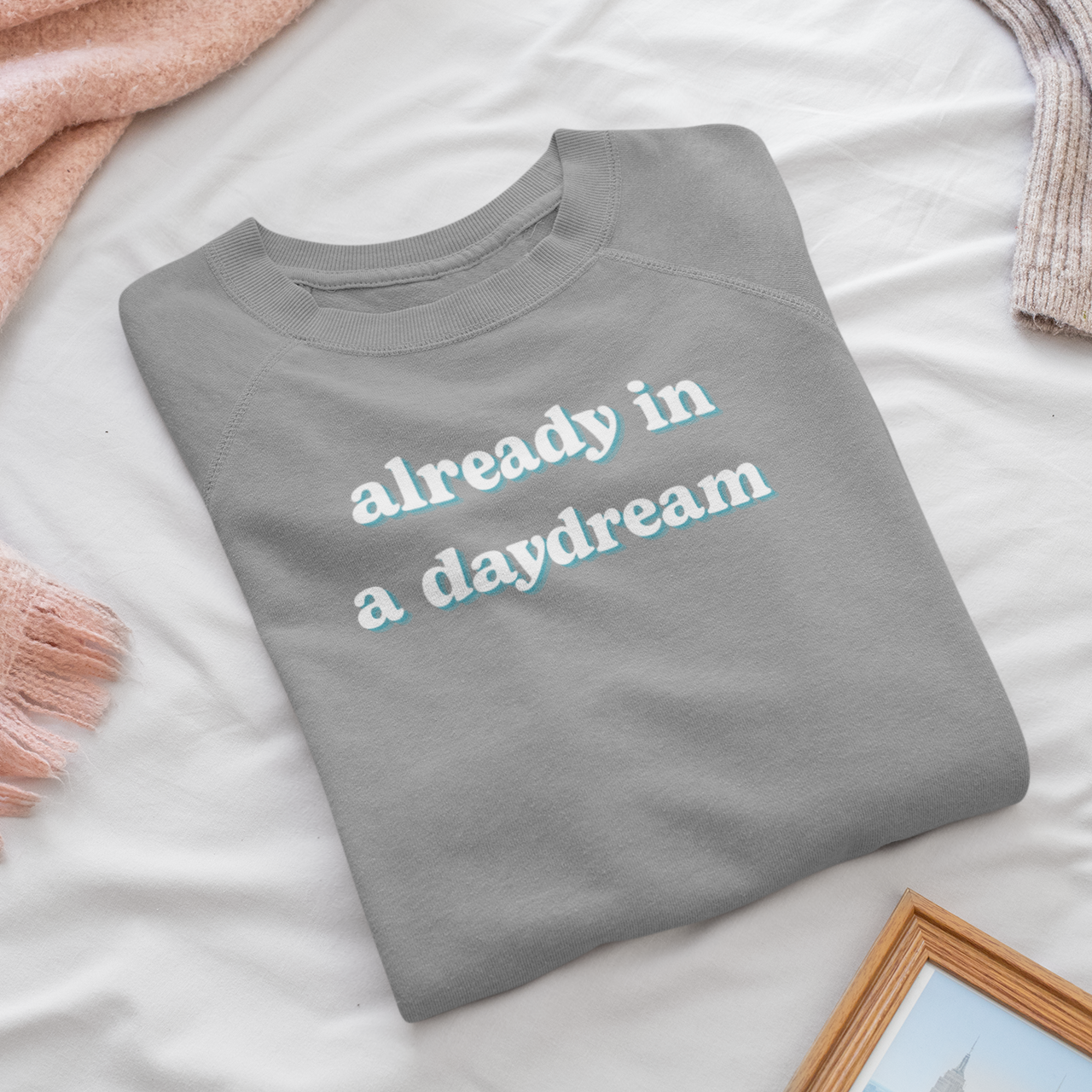 Folded gray sweatshirt with 'already in a daydream' text on a bed with pink blanket and photo frame.