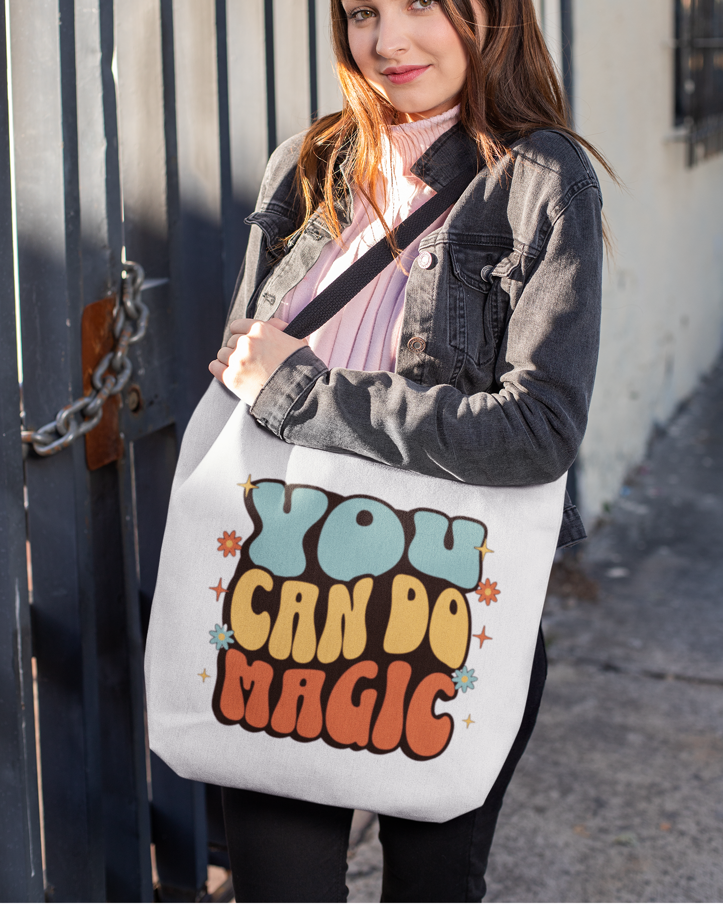Woman holding a tote bag with 'You Can Do Magic' text against a metal gate.
