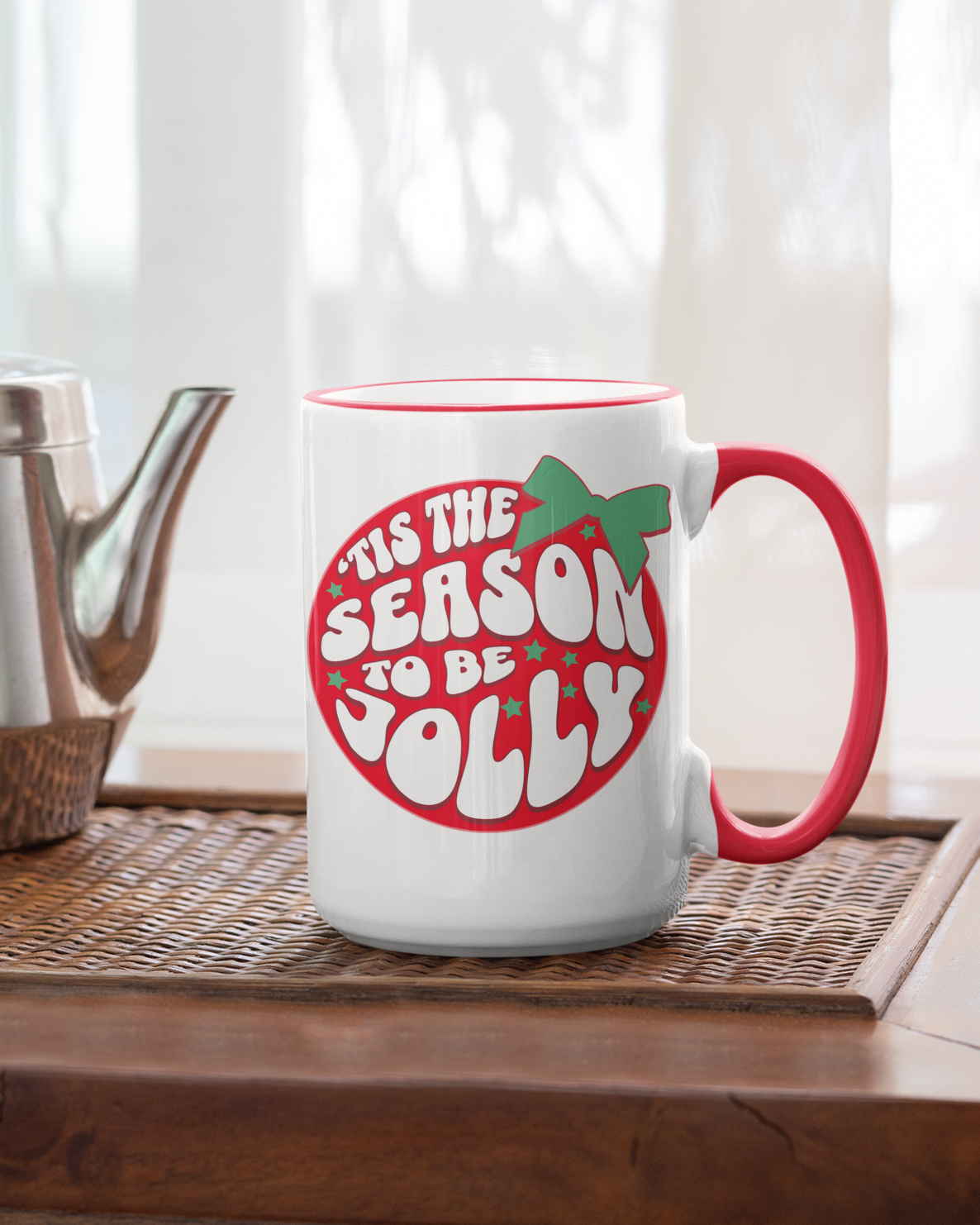 White mug with red handle and 'Tis the Season to be Jolly' text on a wooden surface with a teapot.