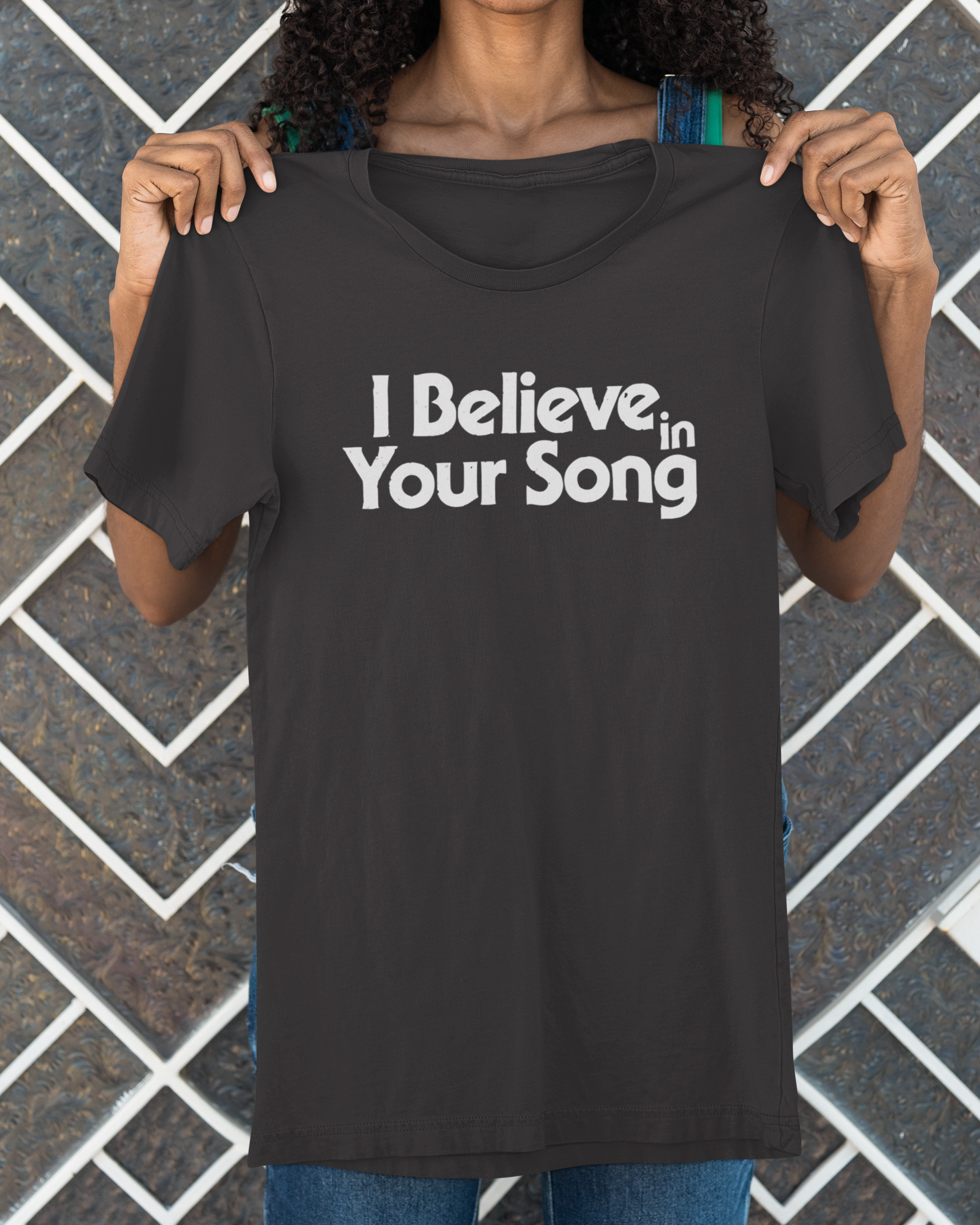 Person wearing a black t-shirt with 'I Believe in Your Song' text against a brick wall.