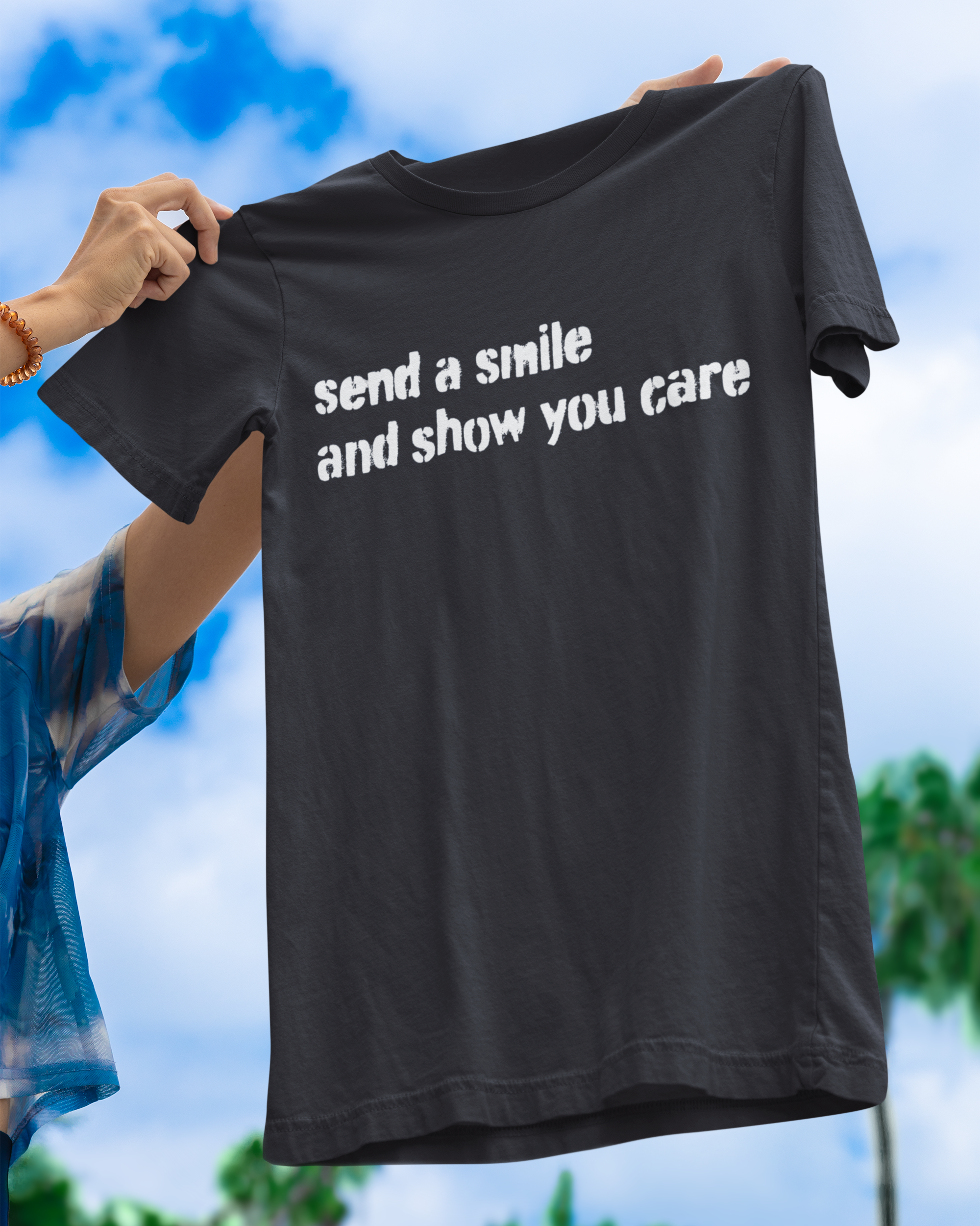 Black t-shirt with 'send a smile and show you care' text held by a person against a blue sky and green trees background.