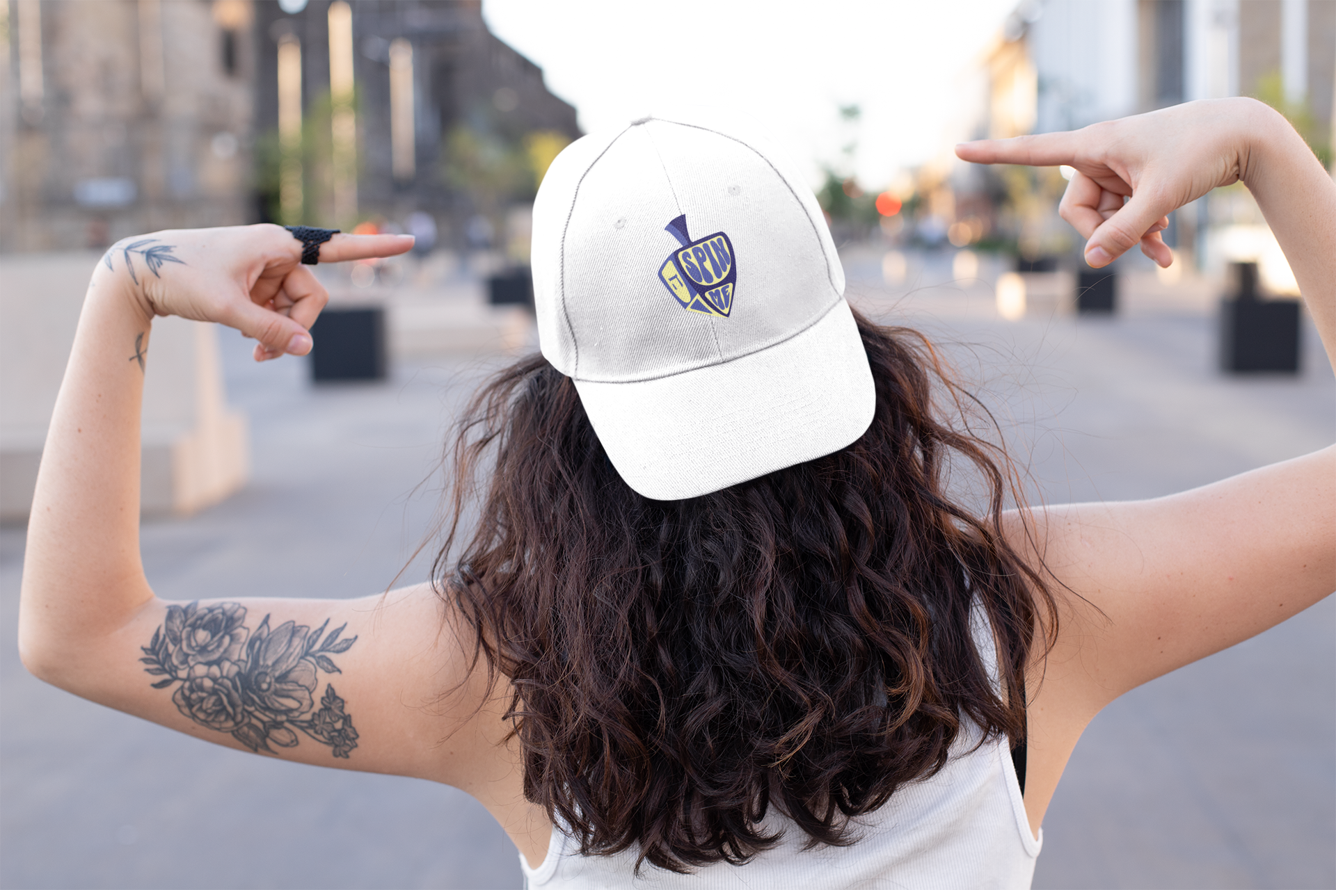 Person wearing a white cap with a logo, pointing towards it, on a blurred street background