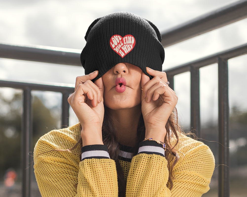 Person wearing a yellow sweater and black cap with a heart design, sitting on a bench.