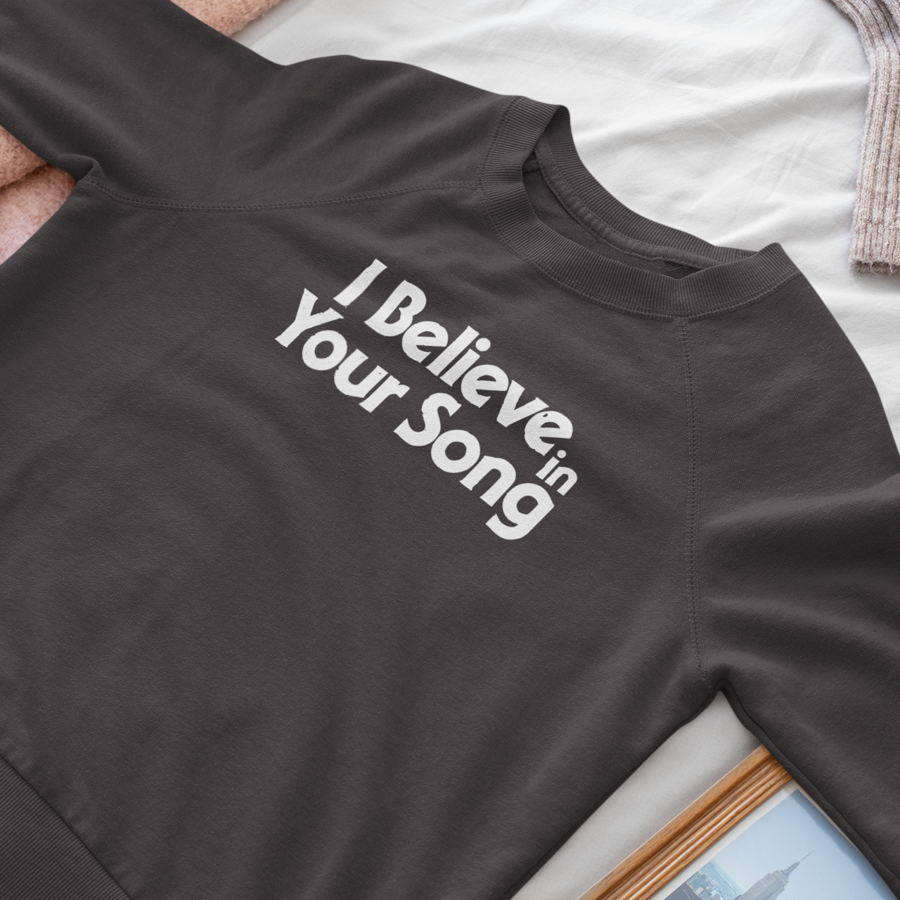 Black sweatshirt with 'I Believe in Your Song' text on a white surface with pink fabric.