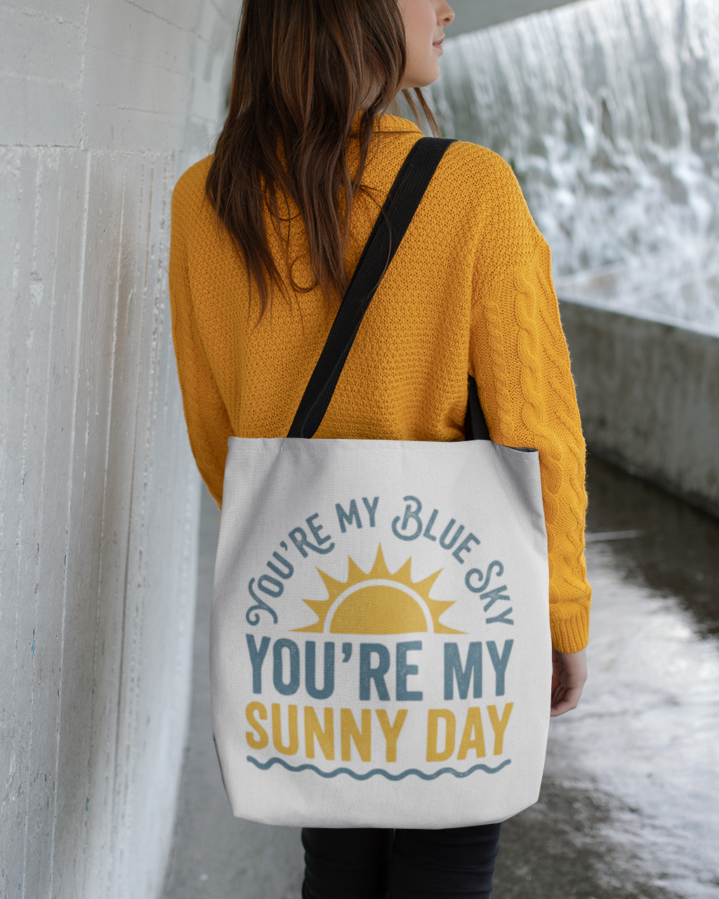 Person holding a tote bag with a motivational quote near a waterfall