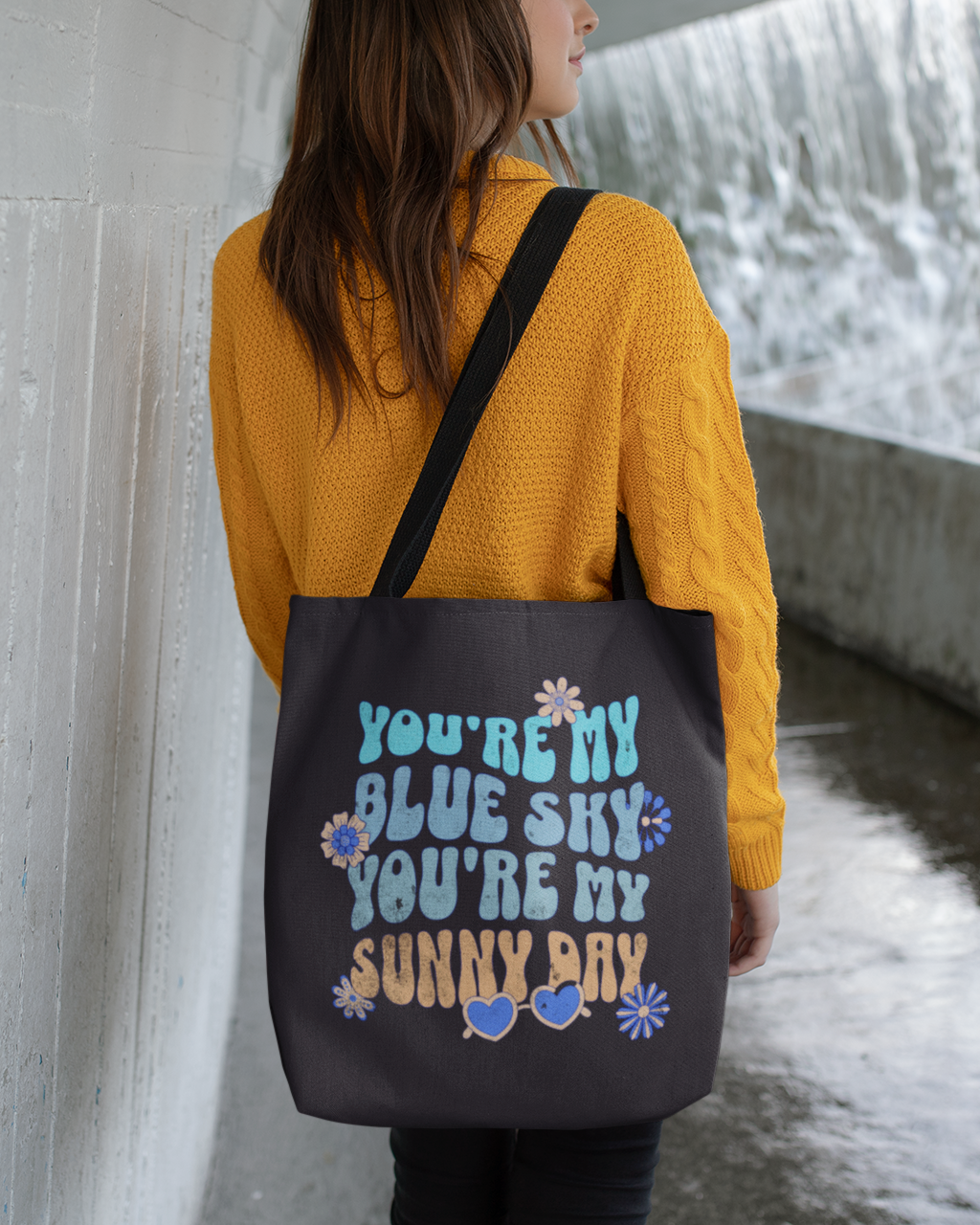 Person wearing a yellow sweater and carrying a black tote bag with text by a water feature