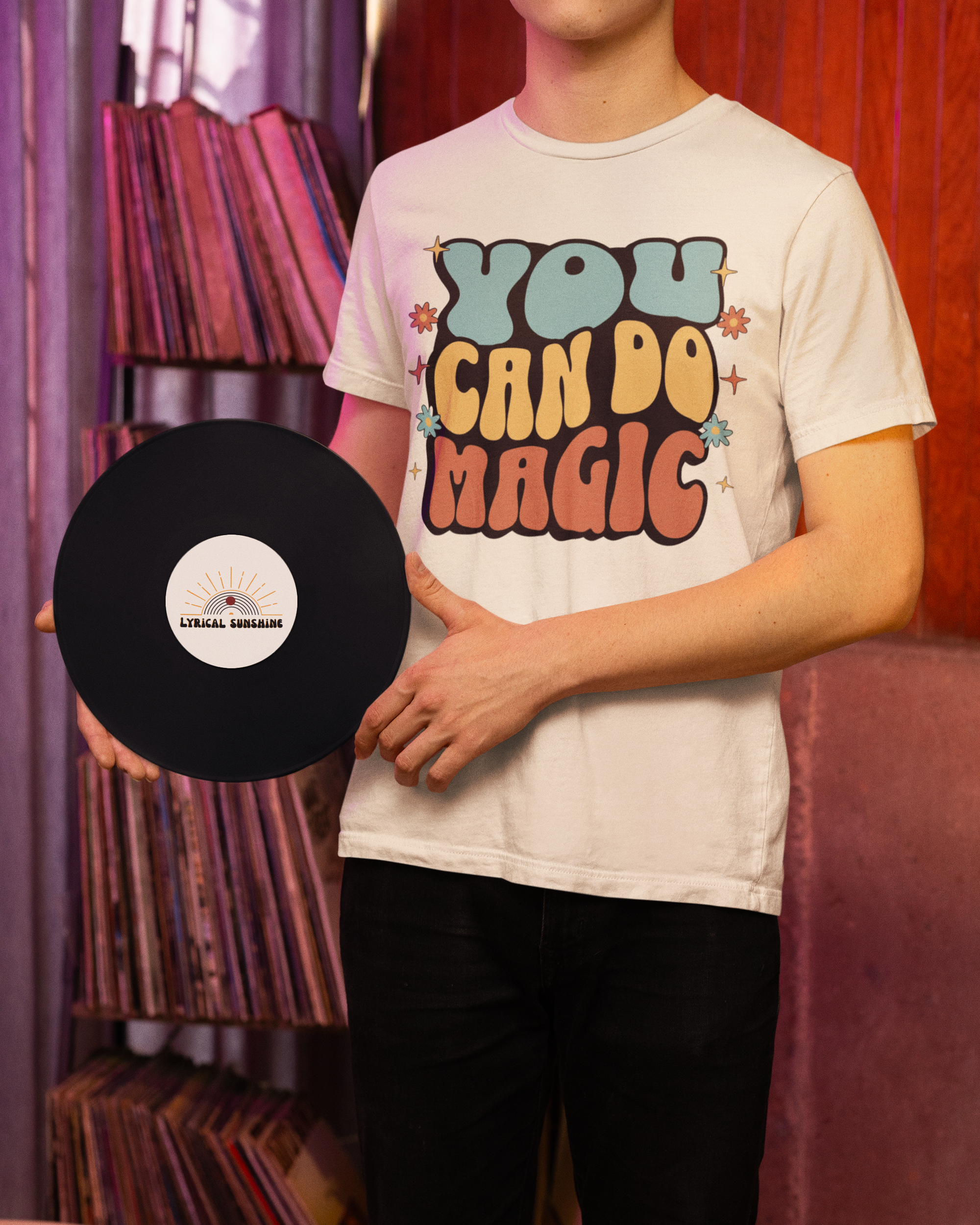 Person wearing a t-shirt with 'You Can Do Magic' text, holding a vinyl record in a room with bookshelves.